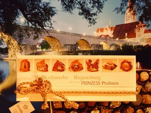 Werbebild des Café Prinzess mit Pralinenschachtel im Vordergrund, im Hintergrund ist die Steinerne Brücke und der Regensburger Dom zu sehen